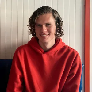 Portrait of Calder. Young white man with medium length light brown hair smiling and wearing a red-orange hoodie.